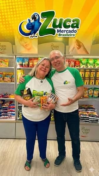 O casal proprietário do Zuca Mercado, de braços dados e a segurar produtos brasileiros, dentro da loja
